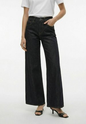 Persoon draagt een zwarte, hooggetailleerde wijde broek met een wit T-shirt en zwarte open teen sandaaltjes met hak, met één hand in de zak.