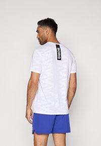 Hvid camouflage-mønstret kortærmet t-shirt med et sort logo-tag på ryggen. Modellen har blå shorts på.