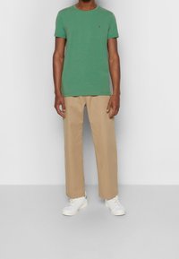 T-shirt en coton vert avec col rond et petit logo, associé à un pantalon droit de couleur crème et des baskets blanches.
