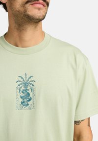 Camiseta de algodón verde claro con un diseño gráfico azul que presenta una serpiente, hojas de palmera y el texto "Balance Opposites RVCA". Estilo de cuello redondo.