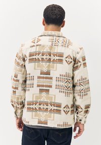 Chemise à motifs beige, présentant des dessins géométriques en marron, gris et rouille. Confectionnée dans un tissu texturé avec des manches longues et des poignets à boutons.