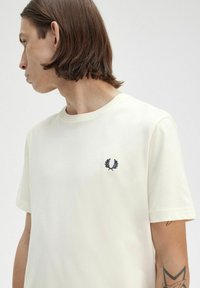 Camiseta corta de manga en color crema, confeccionada en algodón, con un logotipo bordado en navy en el lado izquierdo del pecho. Cuello redondo simple y corte relajado.