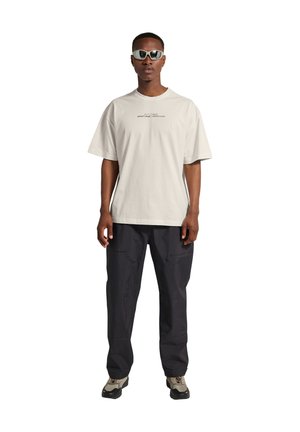 Mann trägt ein weißes, übergroßes T-Shirt mit kleinem Text, schwarze Cargo-Hose, beige und schwarze Sneaker sowie weiße futuristische Sonnenbrillen, steht aufrecht.