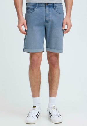 Homme debout portant un short en denim bleu clair retroussé, des chaussettes blanches montantes et des baskets Adidas blanches à rayures noires.