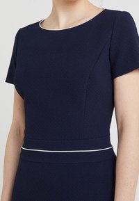 Robe bleu marine texturée à manches courtes, avec un col et une ceinture à bordure blanche, mettant en valeur une silhouette cintrée.
