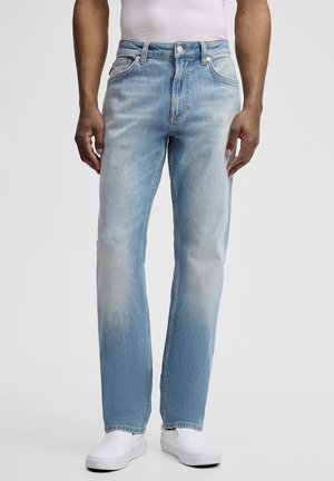 Uomo che indossa jeans azzurri a gamba dritta, scarpe slip-on bianche e una t-shirt bianca infilata, su uno sfondo neutro.