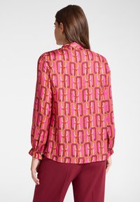 Langarmbluse aus pinkem Satin mit einem geometrischen Muster aus ineinandergreifenden Formen in Orange- und Gelbtönen, mit gerafften Manschetten.