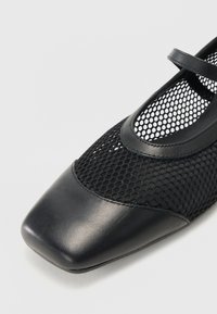 Black shoes featuring a square toe, a combination of smooth leather and mesh material, and a thin strap across the top of the foot.