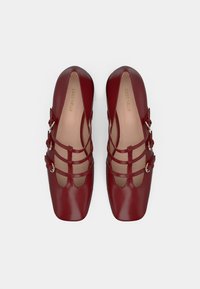 Red leather shoes with a square toe, featuring cut-out designs and multiple straps with silver hardware. Smooth texture and minimalist aesthetic.