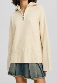 Hellbeige, strukturierter Pullover mit einem 1/2-Reißverschluss-Kragen, geripptem Saum und weiten Ärmeln, über einem Jeans-Minikleid mit fransigem Saum getragen.