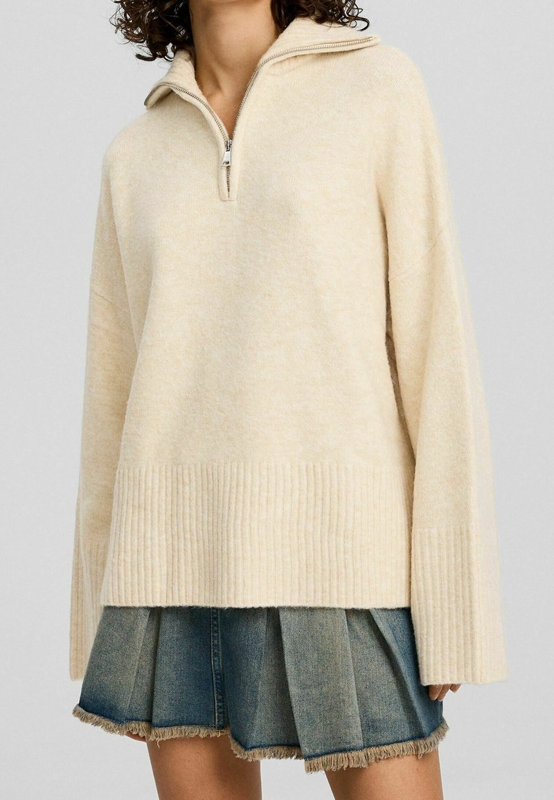 Hellbeige, strukturierter Pullover mit einem 1/2-Reißverschluss-Kragen, geripptem Saum und weiten Ärmeln, über einem Jeans-Minikleid mit fransigem Saum getragen.