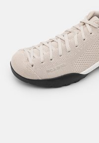 Chaussure de sport beige clair avec une tige tricotée texturée, un bout rond et une semelle extérieure en caoutchouc noir. Comprend des lacets plats et un marquage minimaliste.