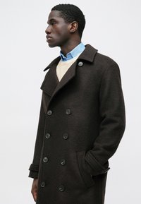 Abrigo marrón de lana con doble botonadura, grandes botones, cuello con muescas y bolsillos laterales, llevado sobre un suéter y camisa de color claro.