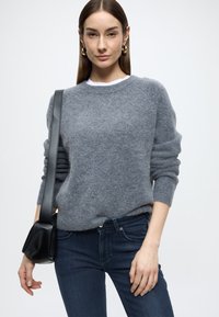 Pelēks vilnas džemperis ar garām piedurknēm un apaļu kaklu, kopā ar tumši ziliem džinsiem un nelielu melnu rokassomu. Gluda tekstūra un ikdienišķa piegriezuma siluets.