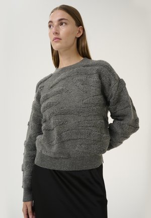 Pull en gris avec une texture et des motifs en relief, col rond, et poignets et ourlet côtelés. Porté par-dessus une jupe noire sur un fond neutre.