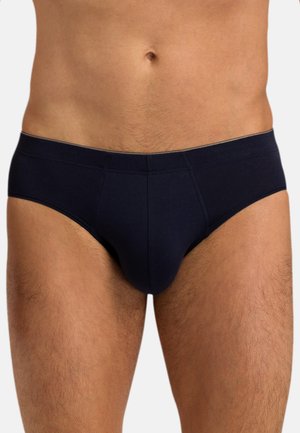 Mann trägt schlichte schwarze Unterwäsche, zeigt den Oberkörper von der Taille bis zu den Oberschenkeln vor einem neutralen Hintergrund.