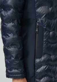 Mornarska puhovka s prešito teksturo in gladkim stranskim materialom. Ima žep z zadrgo. Rahlo sijoča površina s rebrastimi manšetami.