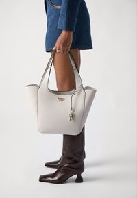 Sac fourre-tout en cuir blanc avec motif texturé, accents argentés, charme en forme de clé et bords foncés contrastants. Modèle portant des bottes marron jusqu'aux genoux.