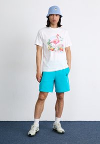 Hvid t-shirt med et farverigt flamingo-grafik, parret med lyse turkise shorts og en lys blå strikket bøttehat. Hvide sneakers og strukturerede sokker.