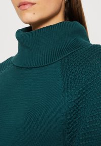 Teal knitted sweater featuring a high, ribbed roll neck and textured pattern on the shoulders. Smooth fabric with a subtle sheen.