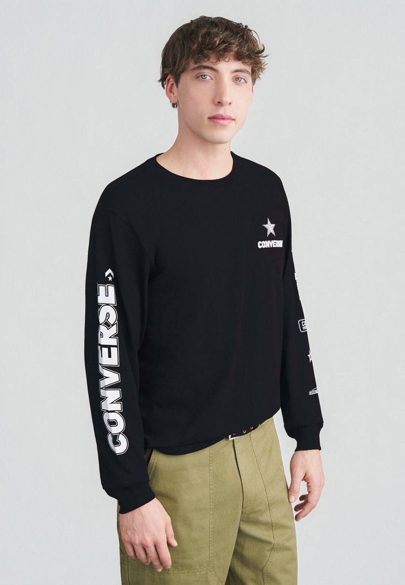 Νεαρός άνδρας που φοράει μαύρο μακρυμάνικο πουκάμισο Converse με λευκό κείμενο λογότυπου στα μανίκια και το στήθος, συνδυασμένο με χακί παντελόνι.