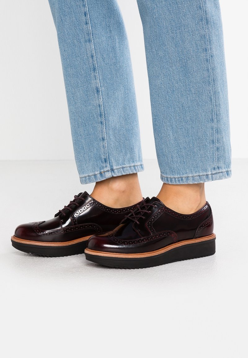 Personne portant des chaussures brogues en cuir bordeaux foncé brillant avec des semelles épaisses noires et un jean bleu clair coupé au-dessus des chevilles.
