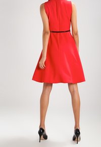 Mujer vestida con un vestido sin mangas de longitud a la rodilla en color rojo, con un cinturón negro y tacones altos negros, de pie con la espalda a la cámara.