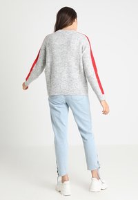 Maglione grigio a coste con strisce rosse lungo le maniche, abbinato a jeans azzurri chiari con lacci laterali e sneakers bianche.