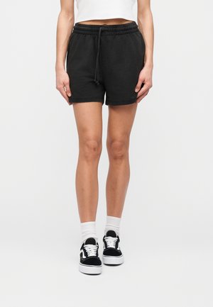 Žena oblečená v čiernych šortkách so sťahovacou šnúrkou, biele ponožky po členky a čierno-biele tenisky na šnurovanie, stojaca na bielom pozadí.