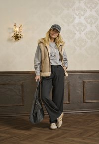Chaleco beige con cuello de piel, sudadera gris con estampado de NYC, pantalones anchos negros, zapatillas ligeras y bolso oscuro, todo frente a una pared con patrón.