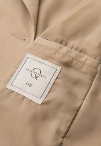Tissu de veste beige avec une étiquette rectangulaire blanche cousue portant l'inscription "Quintessence", taille "L EUF 52", et un logo "Q" stylisé.