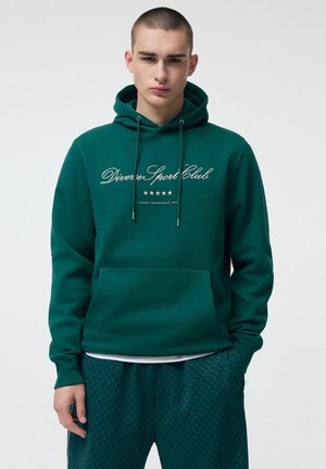 Mężczyzna w ciemnozielonej bluzie z kapturem z napisem „Diverse Sport Club” i dopasowanych wzorzystych spodniach, stojący na tle jednolitego jasnego tła.