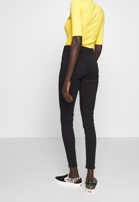 Jeans negros ajustados de cintura alta, combinados con una blusa amarilla de canalé y zapatillas negras sin cordones con un estampado de cebra y detalles de camuflaje.