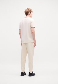 Polo shirt beige clair à manches courtes avec col et accents à rayures foncées, associé à un pantalon de jogging assorti. Des sneakers noires complètent le look.