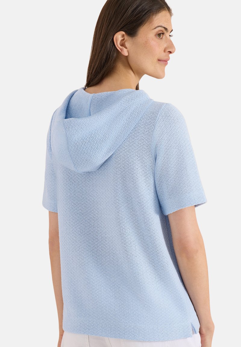 Femme portant un haut en tricot à capuche bleu clair à manches courtes, vue de dos, sur un fond blanc uni.