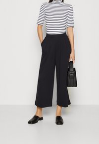 Svarta vida byxor med veck och sidofickor, matchade med en svart och vit randig polotröja och svarta mules, och håller en svart väska.