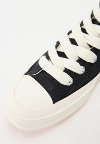 Zapatilla de lona negra con puntera y suela de goma blanca. Cuenta con cordones y ojales blancos, material superior texturizado.