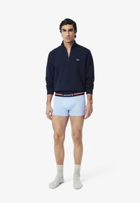 Sweat à zip navy, boxers bleu clair avec ceinture noire, le modèle porte des chaussettes grises, se tenant devant un fond blanc uni.