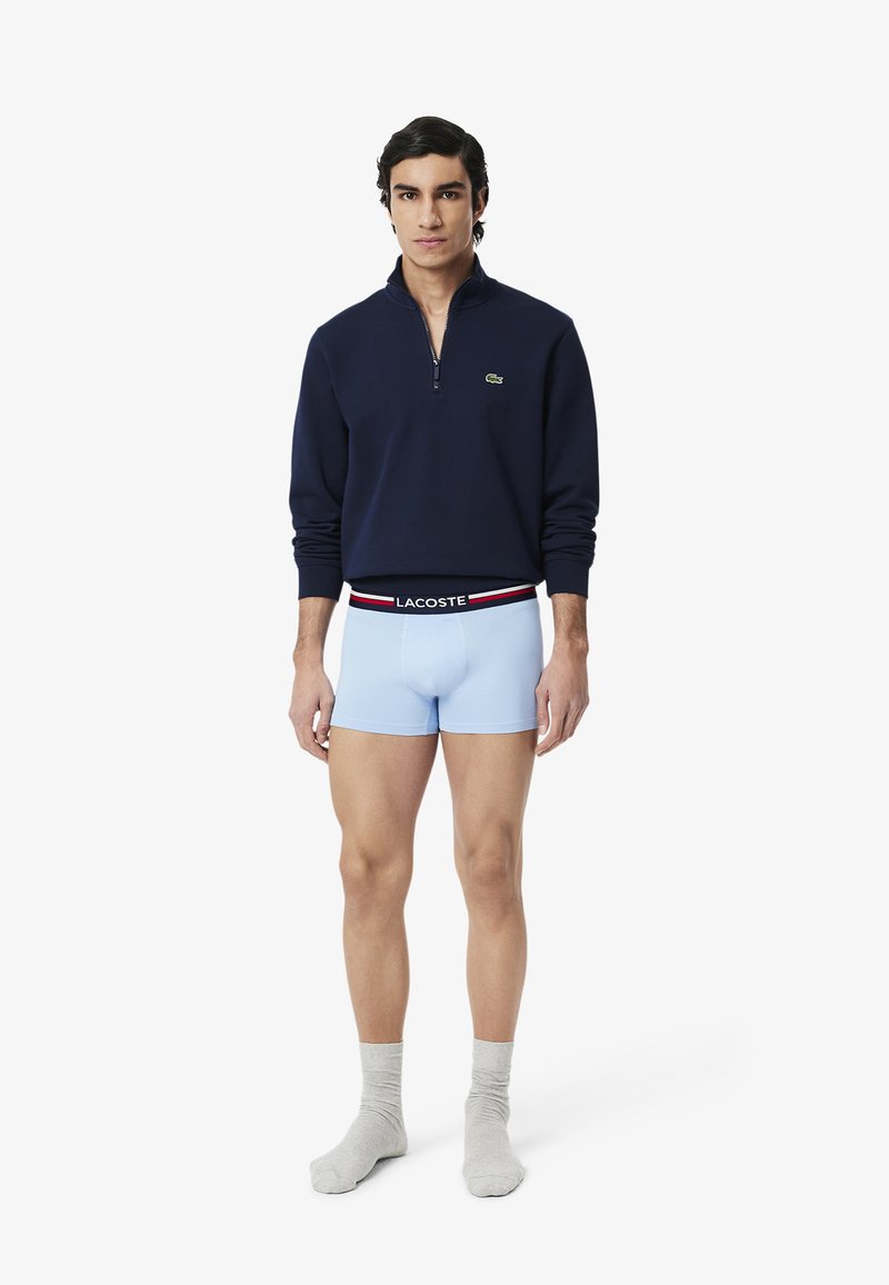 Sweat à zip navy, boxers bleu clair avec ceinture noire, le modèle porte des chaussettes grises, se tenant devant un fond blanc uni.