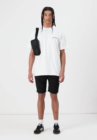 Camiseta de algodón blanca con texto verde, pantalones cortos negros con estampado, bolso bandolera negro y zapatillas negras con acentos blancos, todo sobre un fondo neutro.
