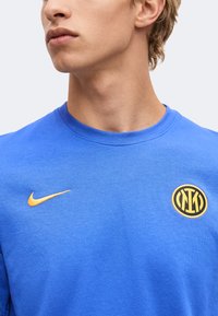 Νεαρό άτομο που φοράει μπλε μπλούζα Nike με κίτρινο λογότυπο swoosh και έμβλημα του ποδοσφαιρικού συλλόγου Ίντερ Μιλάνο στο στήθος.
