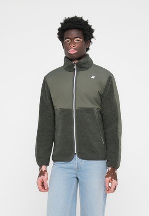 Veste vert olive avec une texture en polaire et un tissu lisse sur les sections supérieures. Fermeture éclair à l'avant et petit logo sur la poitrine.