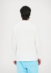 T-shirt bianco a maniche lunghe con scollatura rotonda, tessuto di cotone morbido e dettagli blu sulle maniche. Abbinato a pantaloni azzurri chiari.