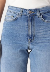Close-up of midsection wearing light blue denim jeans and a white ribbed top, showing button, belt loops, and front pockets.