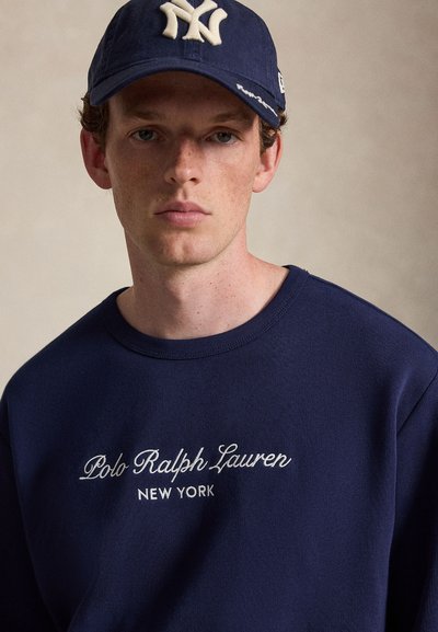 Νέος άντρας που φοράει μπλε σκούρο φούτερ Polo Ralph Lauren και σκούρο μπλε καπέλο μπέιζμπολ των New York Yankees, σε μονόχρωμο φόντο.