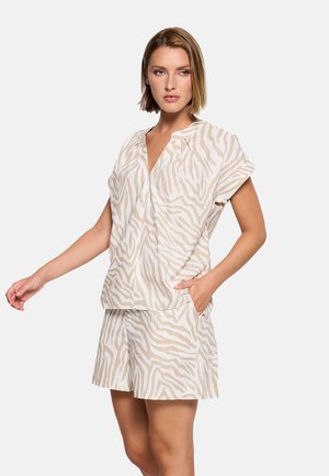 Mujer vestida con top de manga corta a rayas de cebra beige y blanco y pantalones cortos a juego, una mano en el bolsillo, de pie contra un fondo liso.