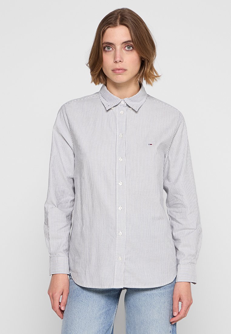 Camicia a maniche lunghe con bottoni, a righe verticali blu e bianche, colletto, e piccolo dettaglio del logo sul petto. Materiale in cotone.