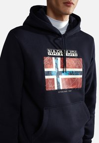 Sudadera azul marino con un bolsillo frontal, que presenta un gráfico de la bandera noruega, acentos blancos y el nombre de la marca "Napapijri". Textura suave.