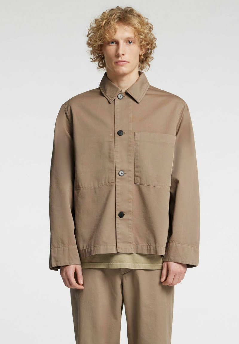 Lys brun jakke laget av glatt stoff, med spiss krage, knappelukking foran, en brystlomme og lange ermer.