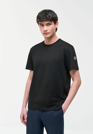 Giovane uomo con capelli corti e scuri che indossa una maglietta nera semplice con una toppa rotonda sul logo nella manica sinistra e pantaloni blu scuro.